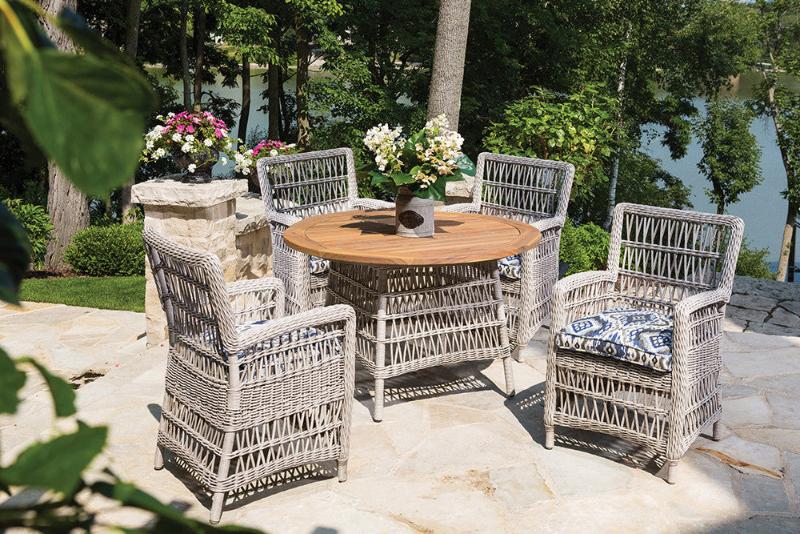 Outdoor patio dining set with a round teak-topped table and four armchairs made of welded aluminum, featuring cushioned seats and backrests in a wicker design.