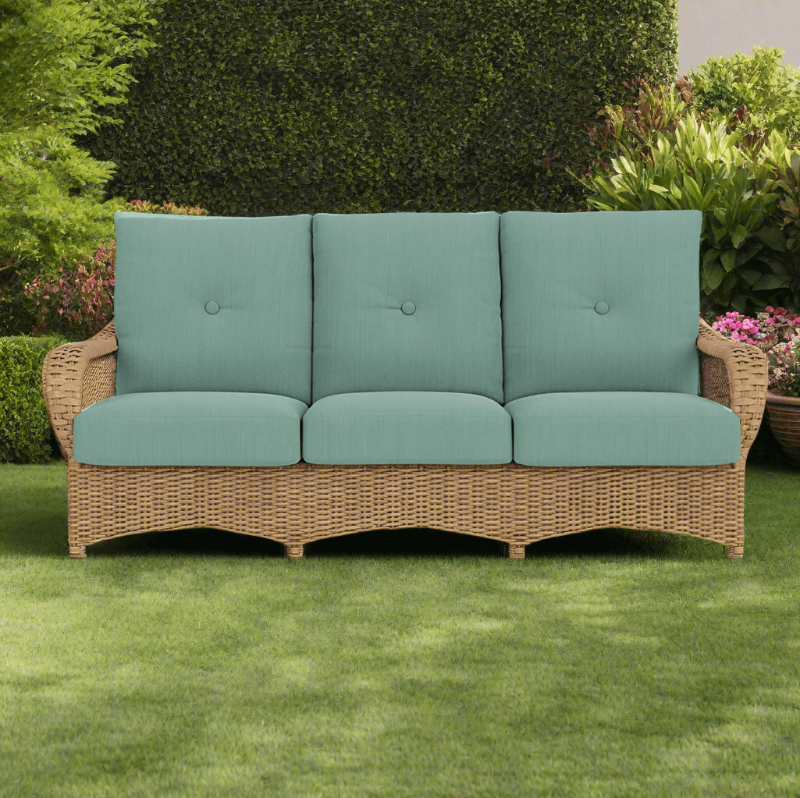 Wicker outdoor sofa with teal cushions on a grassy area with greenery in the background