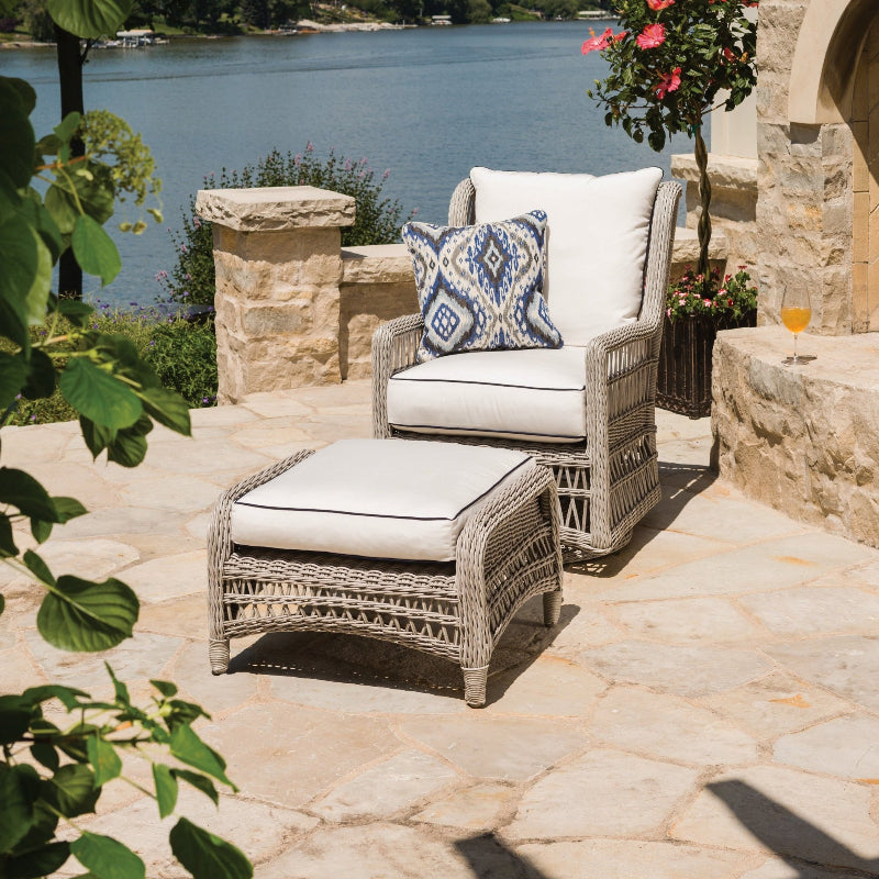 Outdoor patio set with wicker chairs and ottoman on a stone patio by a body of water.