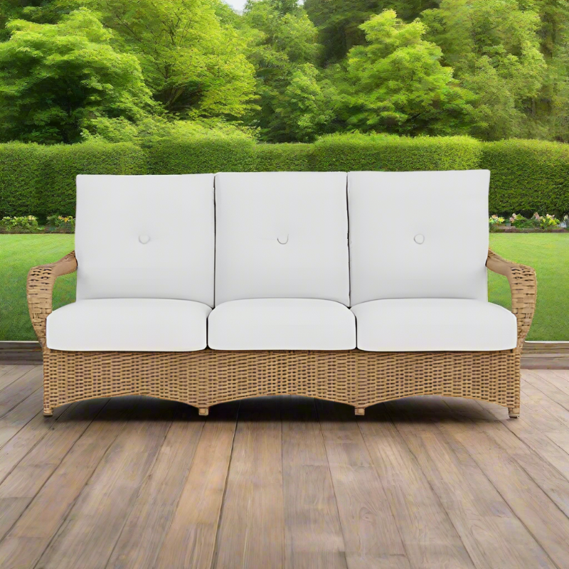 Wicker outdoor sofa with white cushions on a wooden deck with greenery in the background
