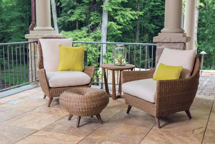 Two wicker chairs with white cushions and yellow pillows on a wooden deck with greenery in the background.