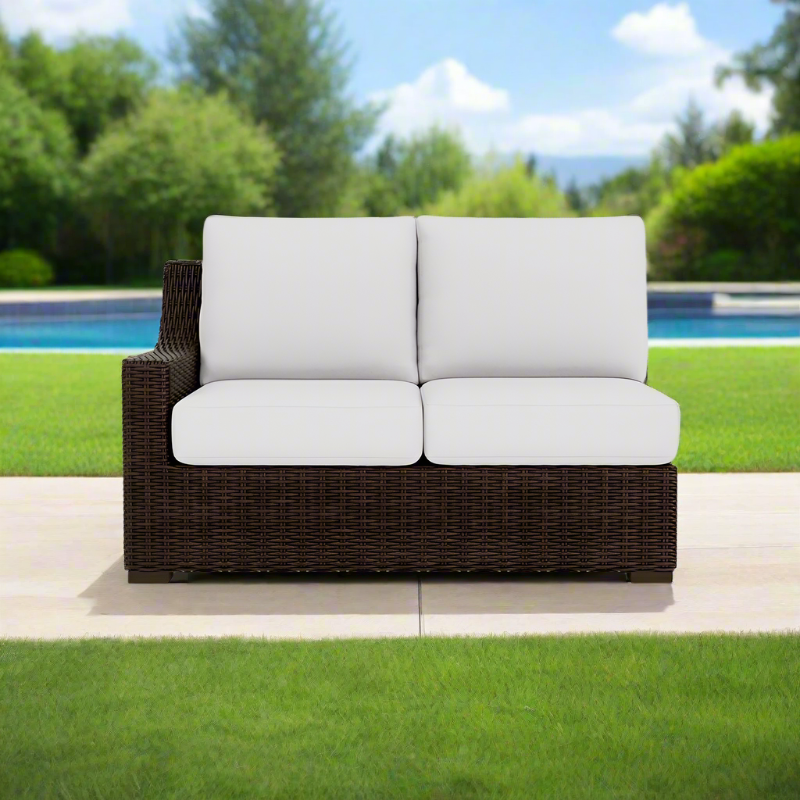 Brown wicker outdoor sofa with white cushions on a patio with a pool and greenery in the background.