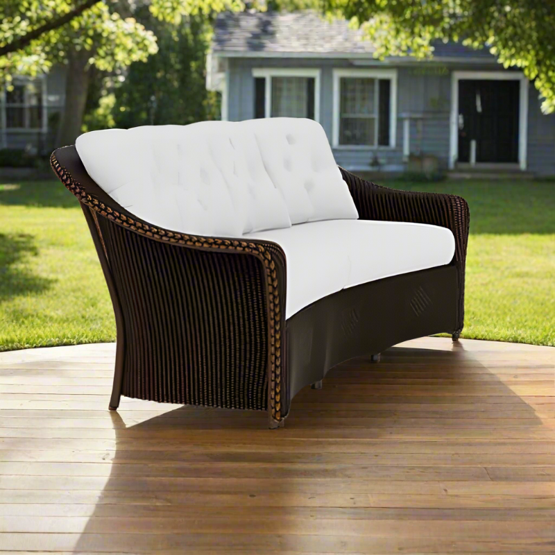 Wicker loveseat with white cushions on a wooden deck in a backyard setting.