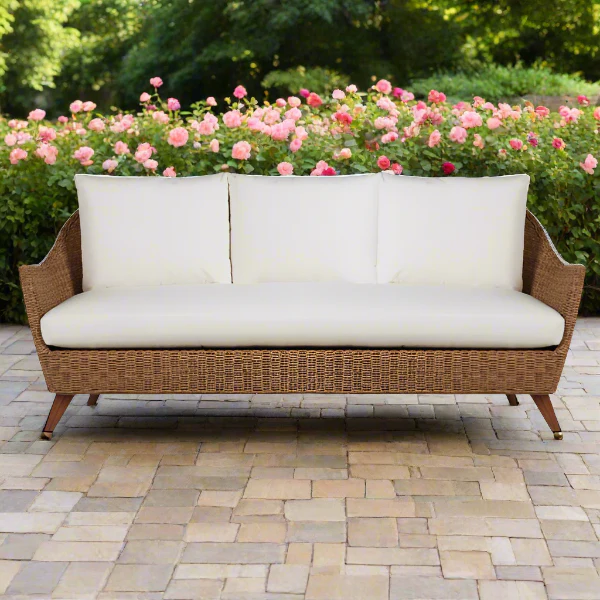 Wicker outdoor sofa with white cushions on a stone patio with pink flowers in the background