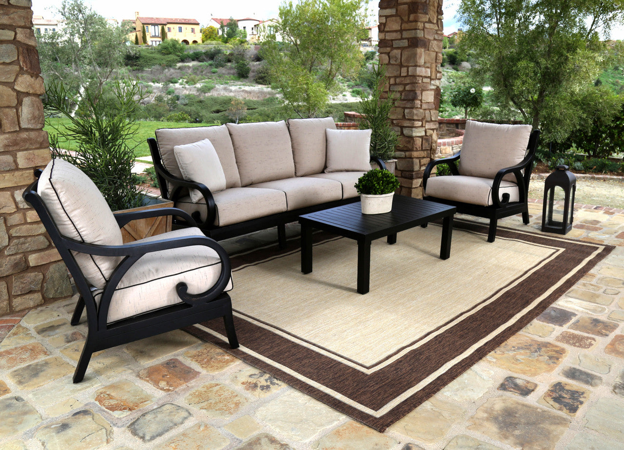 Outdoor furniture set including a sofa, club chairs, and a coffee table made of aluminum with fabric cushions, placed on a patio with a stone wall and shrubbery in the background.