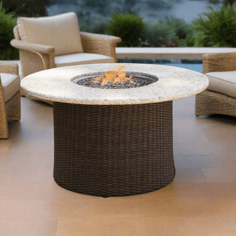Round outdoor fire pit table with wicker design on a patio with chairs and greenery.