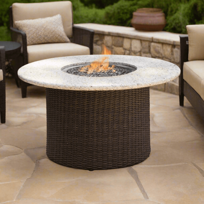 Round outdoor fire pit table with flames on a patio with chairs and a stone wall in the background.