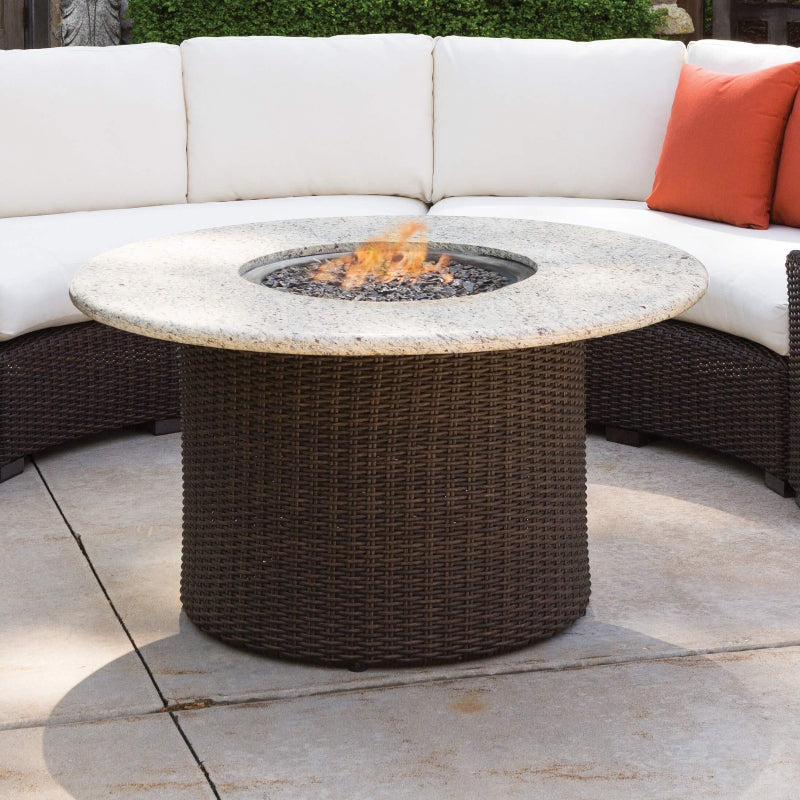 Round fire pit table with flames on a patio, surrounded by a white outdoor sofa with orange cushions.