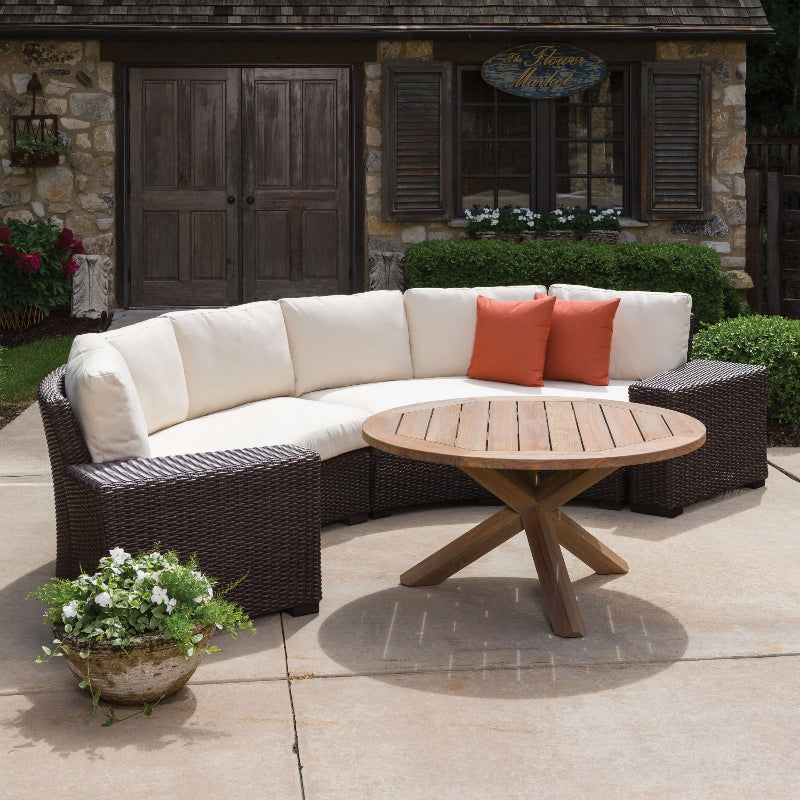 Outdoor patio set with curved sofa, round table, and orange cushions on a stone patio.