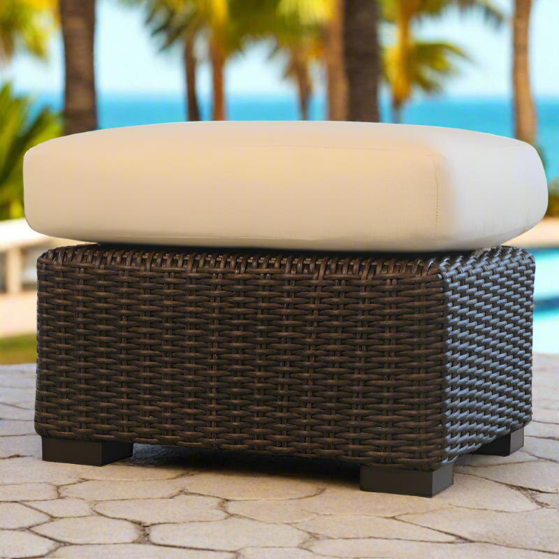 Wicker ottoman with a cream cushion on a patio with palm trees and ocean in the background
