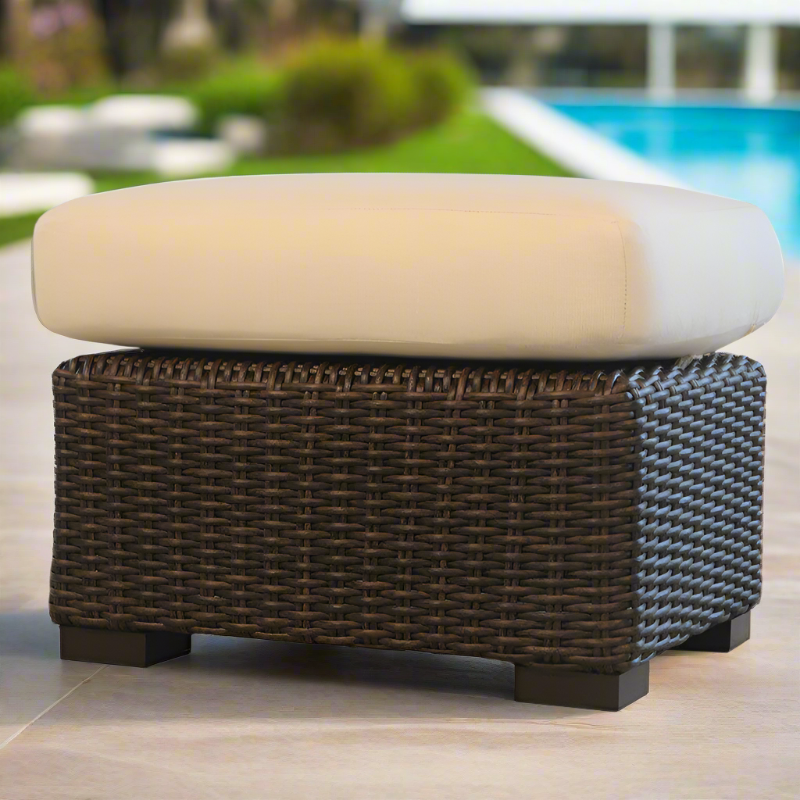 Wicker ottoman with a beige cushion on a patio near a pool
