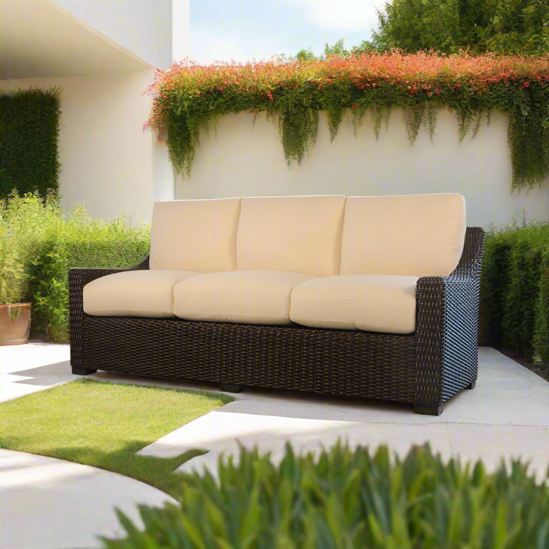 Beige cushioned outdoor sofa with wicker frame on a patio with greenery
