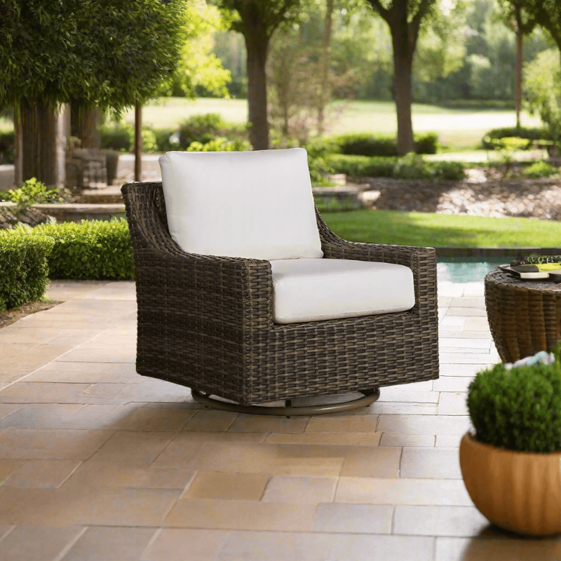 Brown wicker swivel chair with white cushions on a patio with greenery in the background