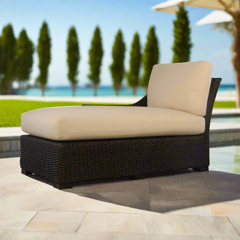 Wicker outdoor lounge chair with beige cushions on a patio with a pool and trees in the background.