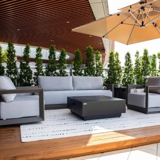Modern outdoor patio furniture set including a light gray three-seater sofa, two matching armchairs, and a dark gray rectangular coffee table on a white patterned rug. The patio features a wooden slatted ceiling, a large beige patio umbrella, and a row of tall green plants in the background.