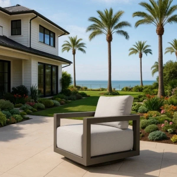 Modern outdoor swivel chair with white cushions in a garden setting with palm trees and a house.