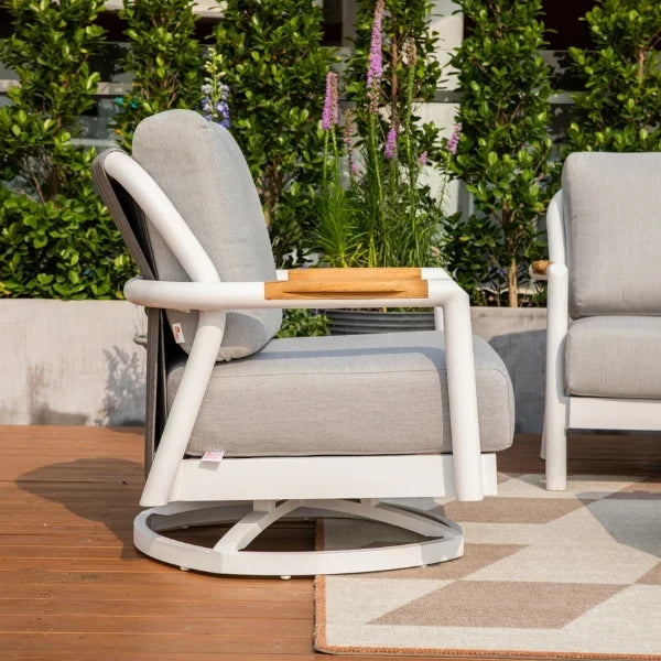 A modern outdoor swivel lounge chair with light gray cushions, a white frame, and a light wood armrest, shown on a wooden deck with green plants in the background.