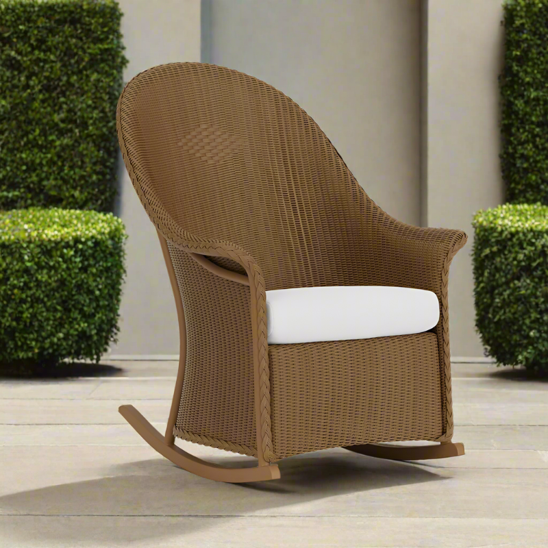 Brown wicker rocking chair with a white cushion on a wooden deck with green bushes in the background.
