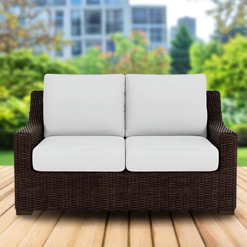 Brown wicker outdoor sofa with white cushions on a wooden deck with a blurred green garden background