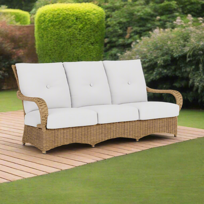 Wicker outdoor sofa with white cushions on a wooden deck with greenery in the background