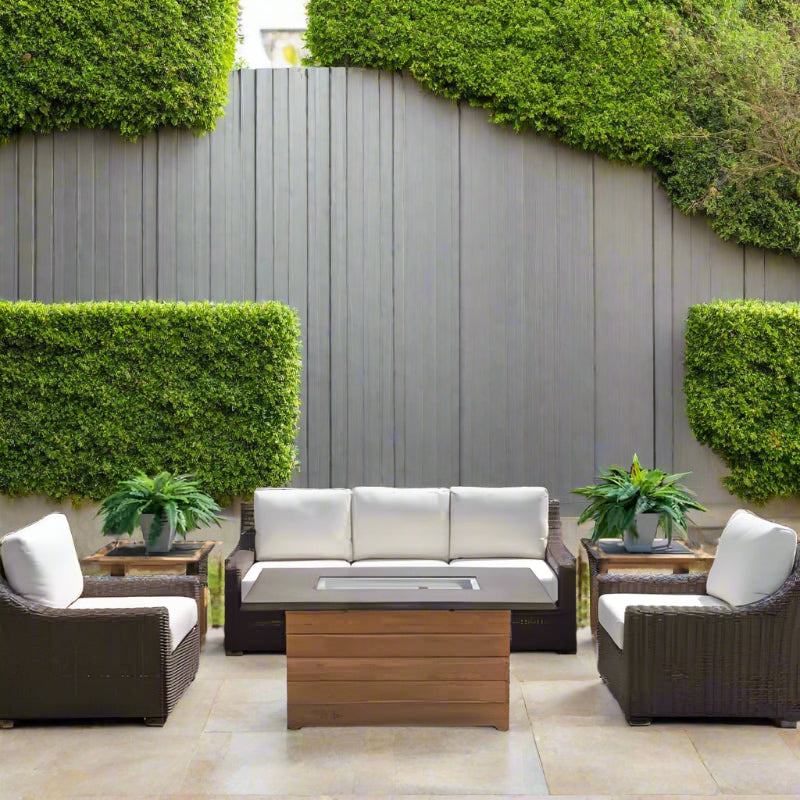 Outdoor patio set with a fire pit table, chairs, and a sofa against a gray wall with greenery.