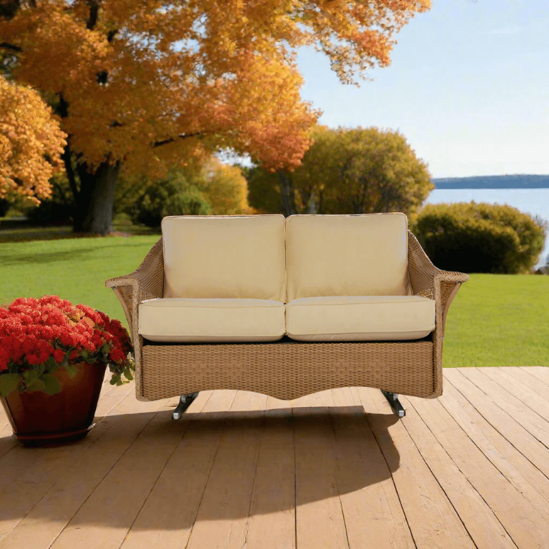 Wicker outdoor loveseat with beige cushions on a wooden deck with autumn trees and flowers in the background.